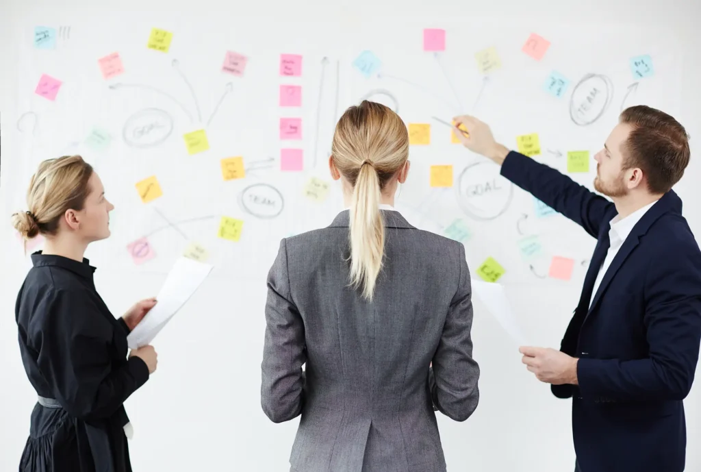 Two businesswomen looking at whiteboard while their colleague explaining scheme of teamwork