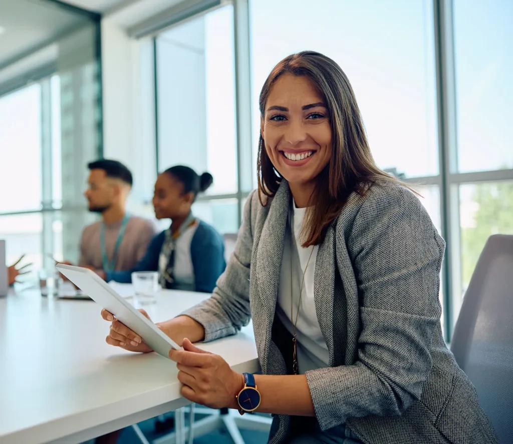 Young happy businesswoman using touchpad during meeting in office and looking at camera.